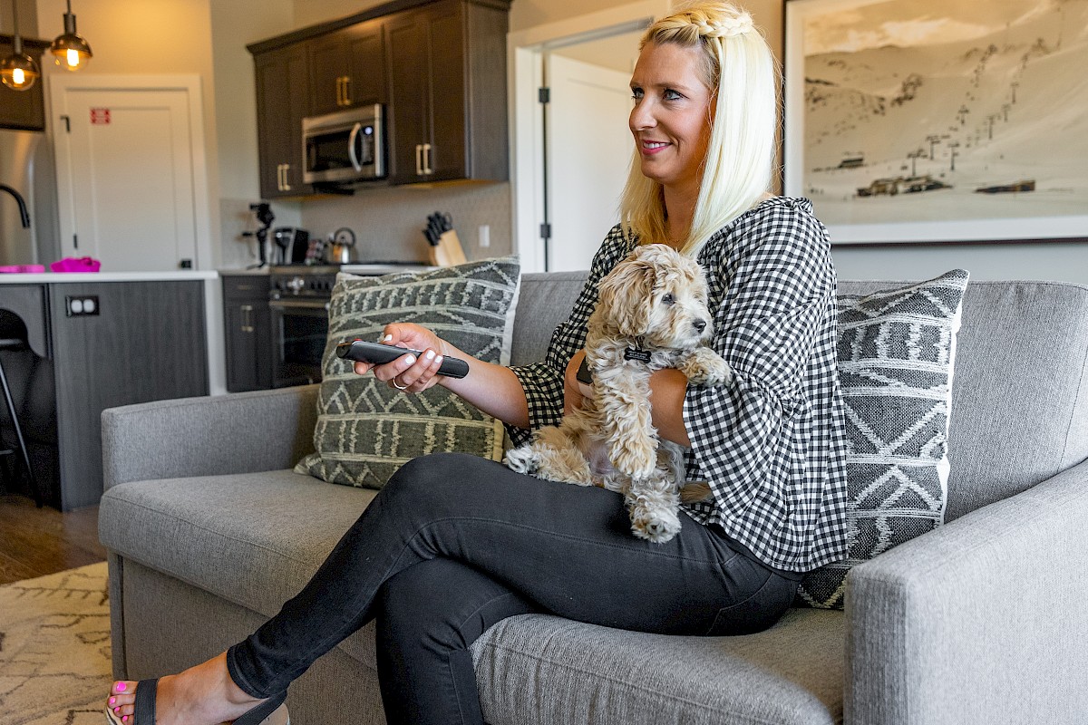 A person sits on a couch with a dog, holding a remote. The background shows a modern kitchen and a large wall photo.