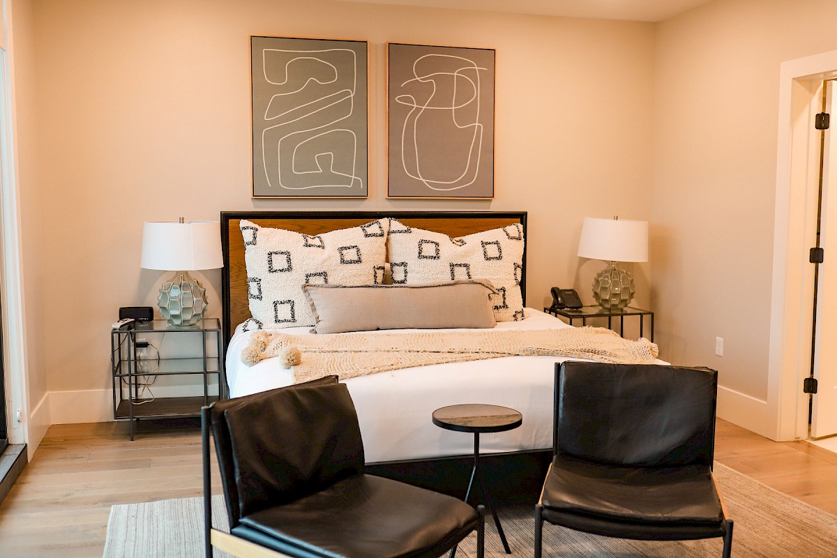 A modern bedroom with a bed, abstract artwork, lamps, and two black chairs facing a small table on a light wood floor.