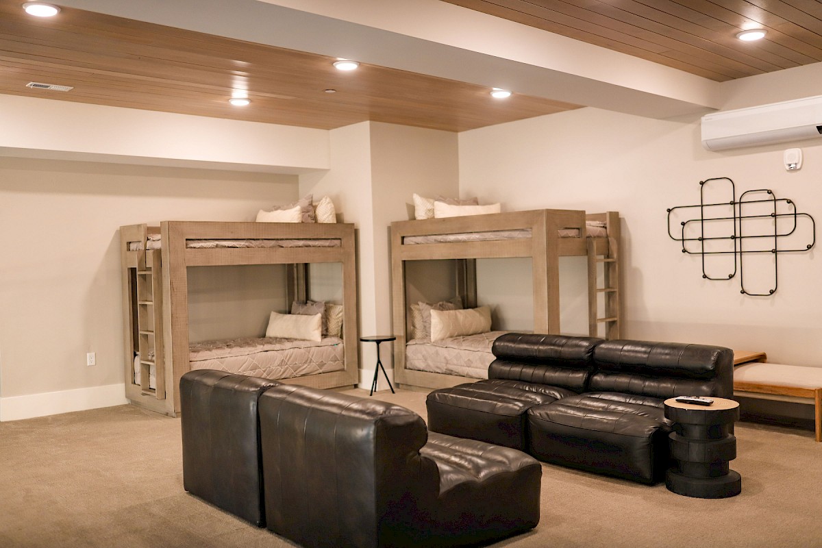 The image shows a room with two sets of bunk beds, a sectional sofa, a wooden ceiling, and modern wall decor.