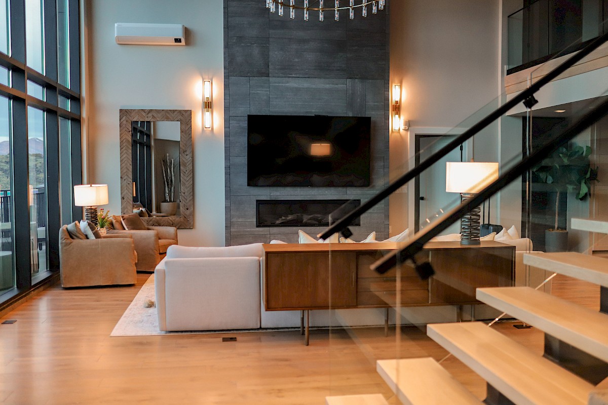 A modern living room with large windows, a sectional sofa, TV, and a staircase with glass railings. Neutral tones and ambient lighting.