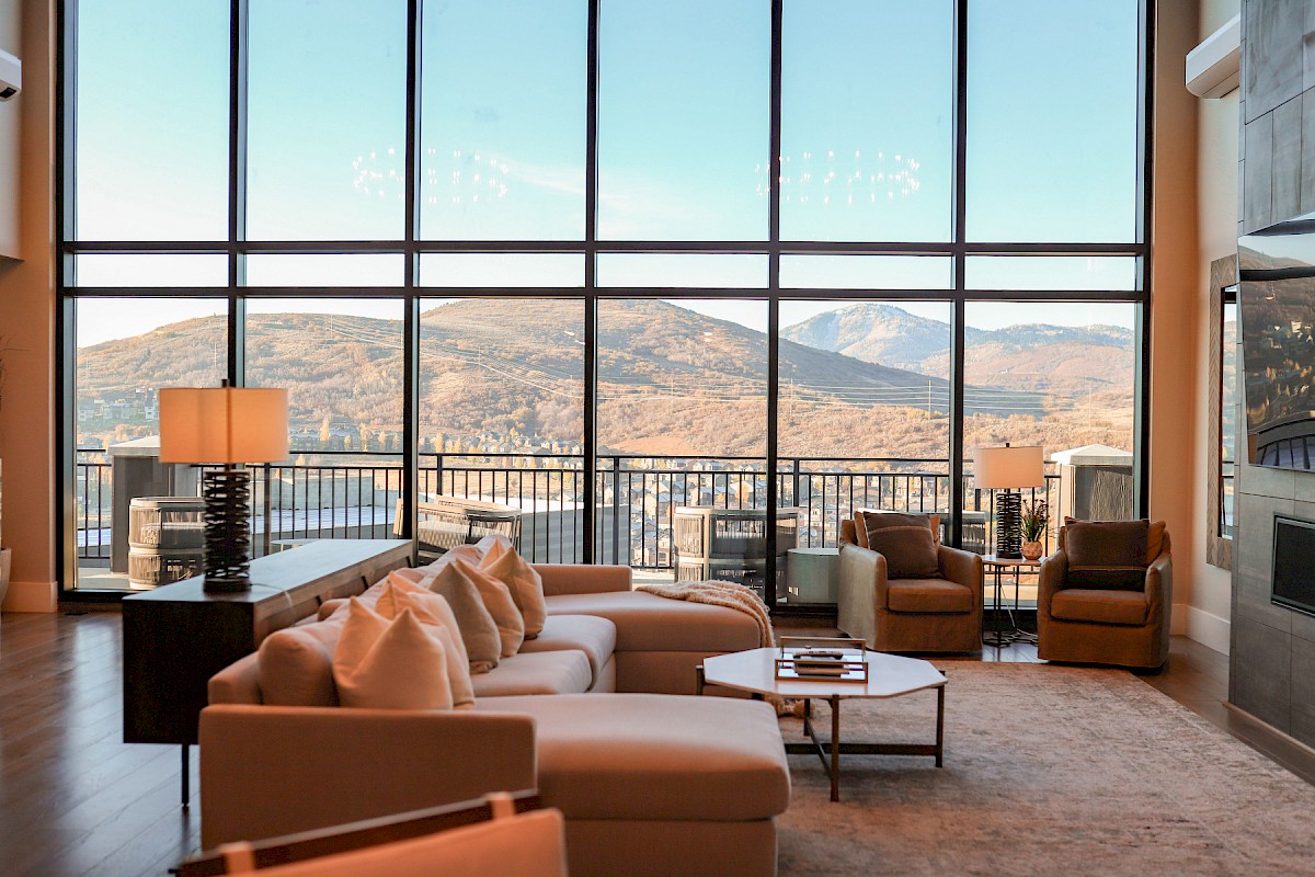 A modern living room with large windows showcasing a scenic mountain view, featuring cozy seating, lamps, and a central coffee table.