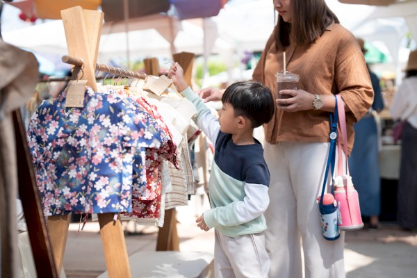 A child and adult browse colorful clothing at an outdoor market. The adult holds a drink and has a bag with a bottle attached.