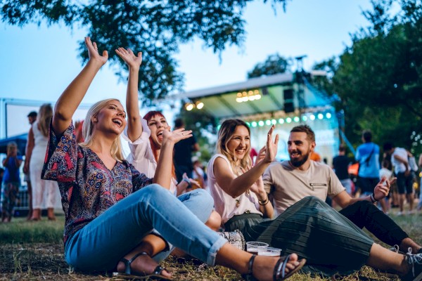 A group of people sitting outdoors, enjoying a lively event with a stage and lights in the background.