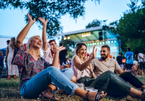 A group of people sit on the grass, enjoying a festival atmosphere with raised hands and smiles, stage lights visible in the background.