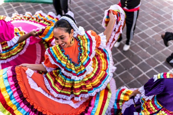 A person is dancing in a colorful, traditional Mexican dress with vibrant ruffles, smiling, and surrounded by more dancers.