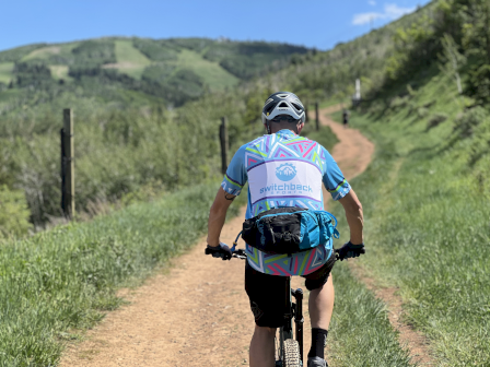 A cyclist is riding on a dirt path through a scenic, grassy landscape with hills in the background under a clear blue sky.