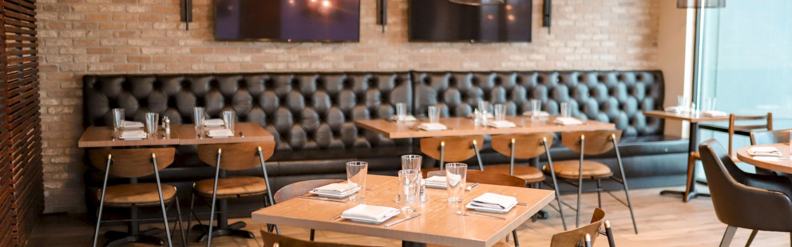 The image shows a cozy restaurant interior with wooden tables, leather chairs, a brick wall, and wall-mounted TVs.