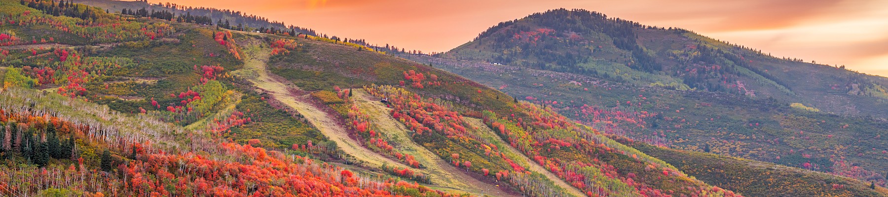 A town lies below colorful, autumnal hills with vibrant orange and red foliage under a dramatic, cloudy sunset sky.