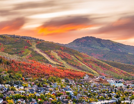 A town lies below colorful, autumnal hills with vibrant orange and red foliage under a dramatic, cloudy sunset sky.