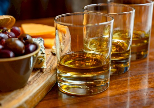Three glasses of whiskey on a wooden table alongside a cheese and grape platter.