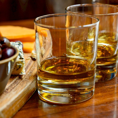 Three glasses of whiskey on a wooden table alongside a cheese and grape platter.