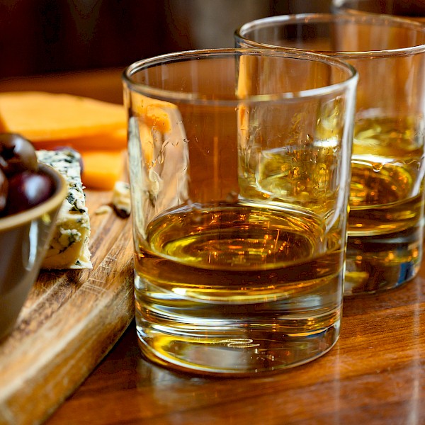 Three glasses of whiskey on a wooden table alongside a cheese and grape platter.