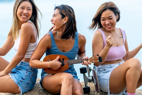 Three people are sitting outdoors, smiling. One holds a ukulele, while all wear casual summer clothes and appear to be enjoying their time.