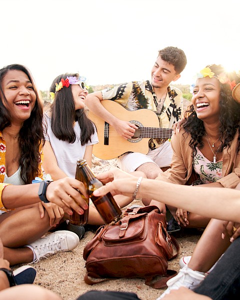 A group of friends sits outdoors, laughing and chatting while one plays the guitar. They appear to be enjoying a relaxed and joyful moment.