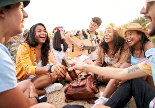 A group of friends sits outdoors, laughing and chatting while one plays the guitar. They appear to be enjoying a relaxed and joyful moment.