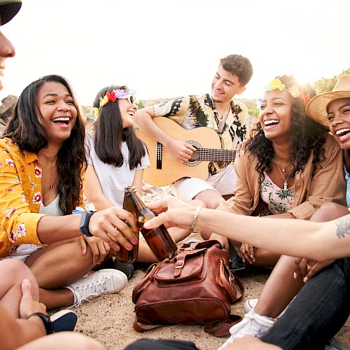 A group of friends sits outdoors, laughing and chatting while one plays the guitar. They appear to be enjoying a relaxed and joyful moment.