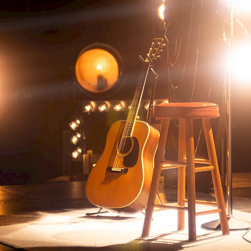 A wooden guitar leans against a stool on stage, surrounded by bright studio lights, creating a warm, inviting atmosphere.