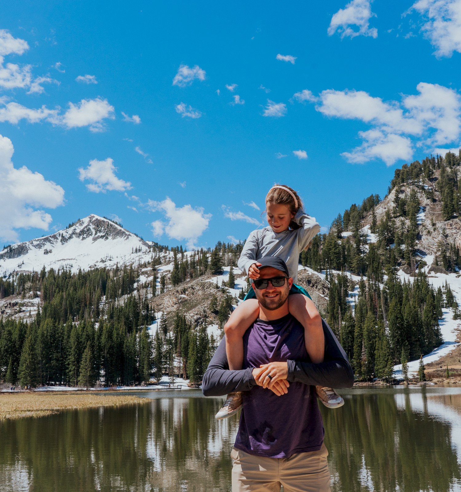 A person carries a child on their shoulders near a lake with snow-capped mountains and trees in the background under a blue sky.