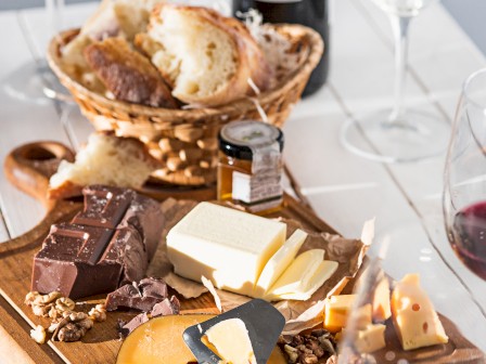 A wooden board with various cheeses, nuts, and chocolate sits on a table with wine glasses, a wine bottle, and a basket of bread.
