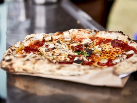 A wood-fired pizza topped with pepperoni, mushrooms, and cheese on a paper sheet, positioned on a metal surface.