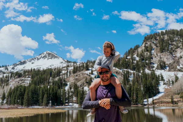 A person carries a child on their shoulders by a scenic lake with snow-capped mountains and a clear blue sky in the background.