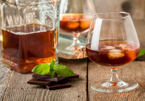 A glass of amber-colored liquor with ice, a bottle, dark chocolate pieces, and mint leaves on a wooden surface.