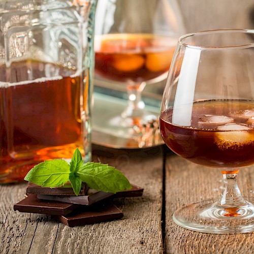 A glass of amber-colored liquor with ice, a bottle, dark chocolate pieces, and mint leaves on a wooden surface.