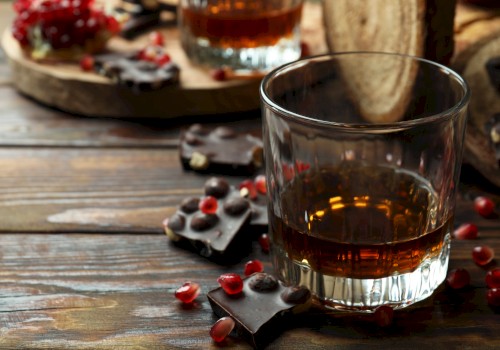 A glass of dark liquor with chocolate pieces and pomegranate seeds on a wooden surface.