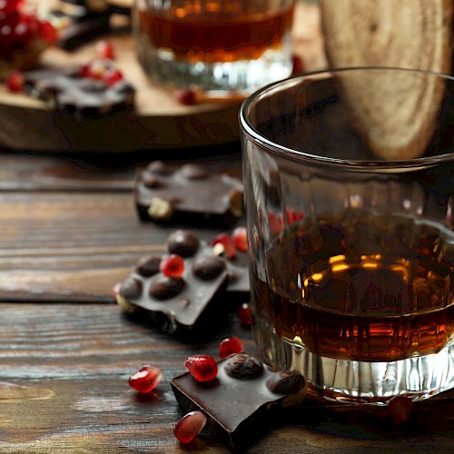 A glass of dark liquor with chocolate pieces and pomegranate seeds on a wooden surface.