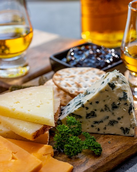 A wooden board with assorted cheeses, crackers, a garnish, and two glasses of whiskey.