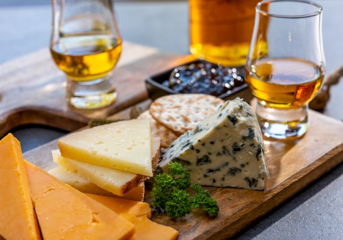A wooden board with assorted cheeses, crackers, a garnish, and two glasses of whiskey.