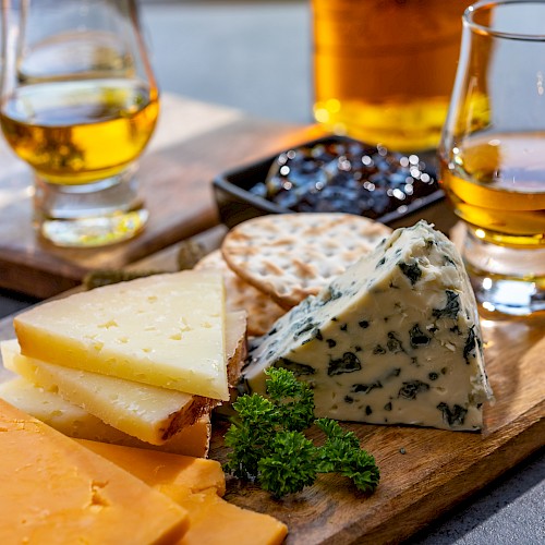 A wooden board with assorted cheeses, crackers, a garnish, and two glasses of whiskey.