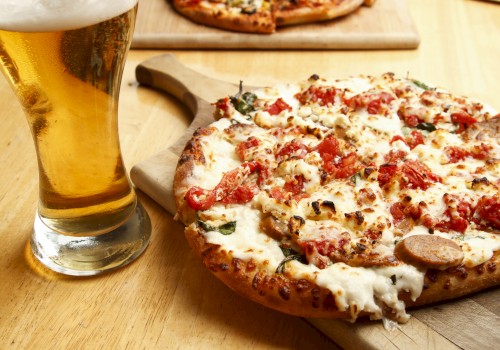 A glass of beer sits next to a cheesy, tomato-topped pizza on a wooden board, set on a table.