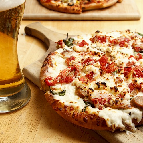 A glass of beer sits next to a cheesy, tomato-topped pizza on a wooden board, set on a table.
