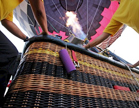 A hot air balloon being prepared with a burner flame, visible basket, two people in yellow shirts handling it, and colorful balloon canopy.