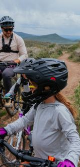 Two people, wearing helmets and biking gear, pause on mountain bikes on a dirt trail surrounded by greenery and hills under a cloudy sky.