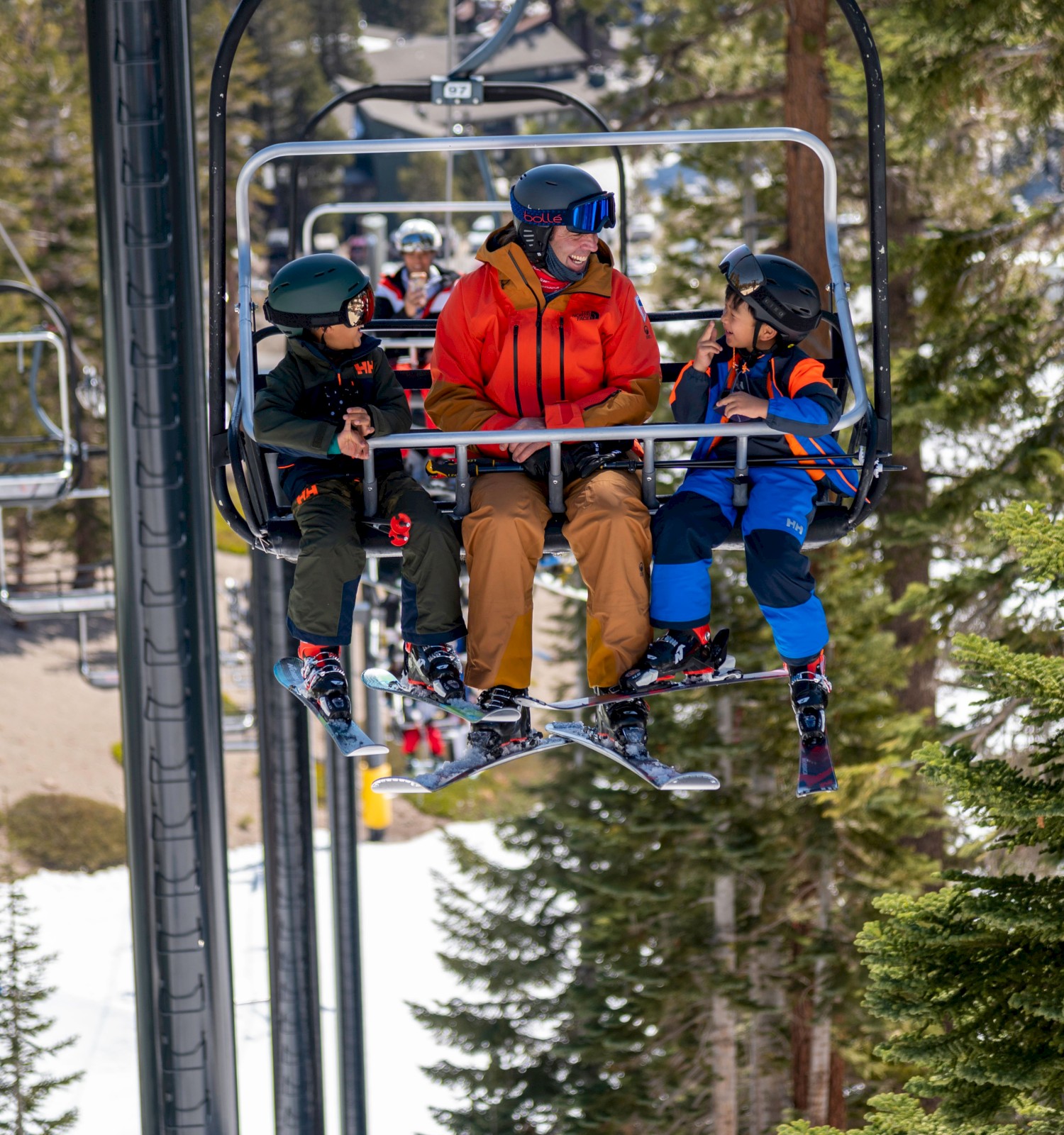 Three people are sitting on a ski lift, surrounded by snow and trees, wearing winter gear and helmets, enjoying a day on the slopes.