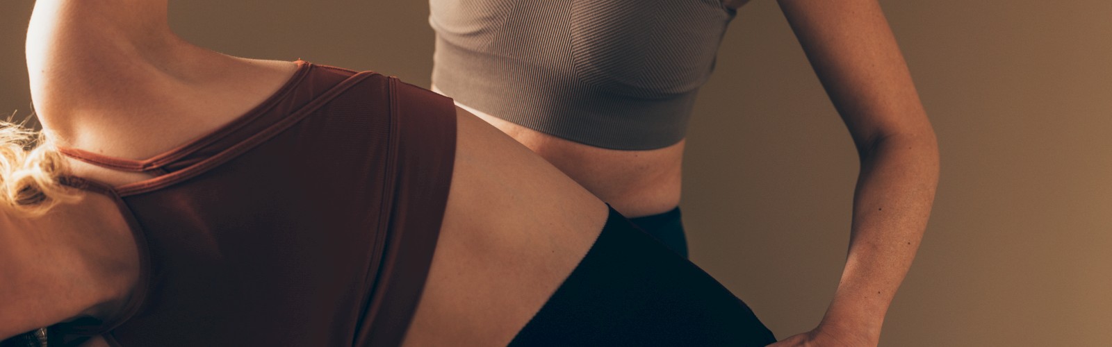 Two people are engaged in a yoga or stretching pose, wearing sports bras and leggings, against a neutral background.