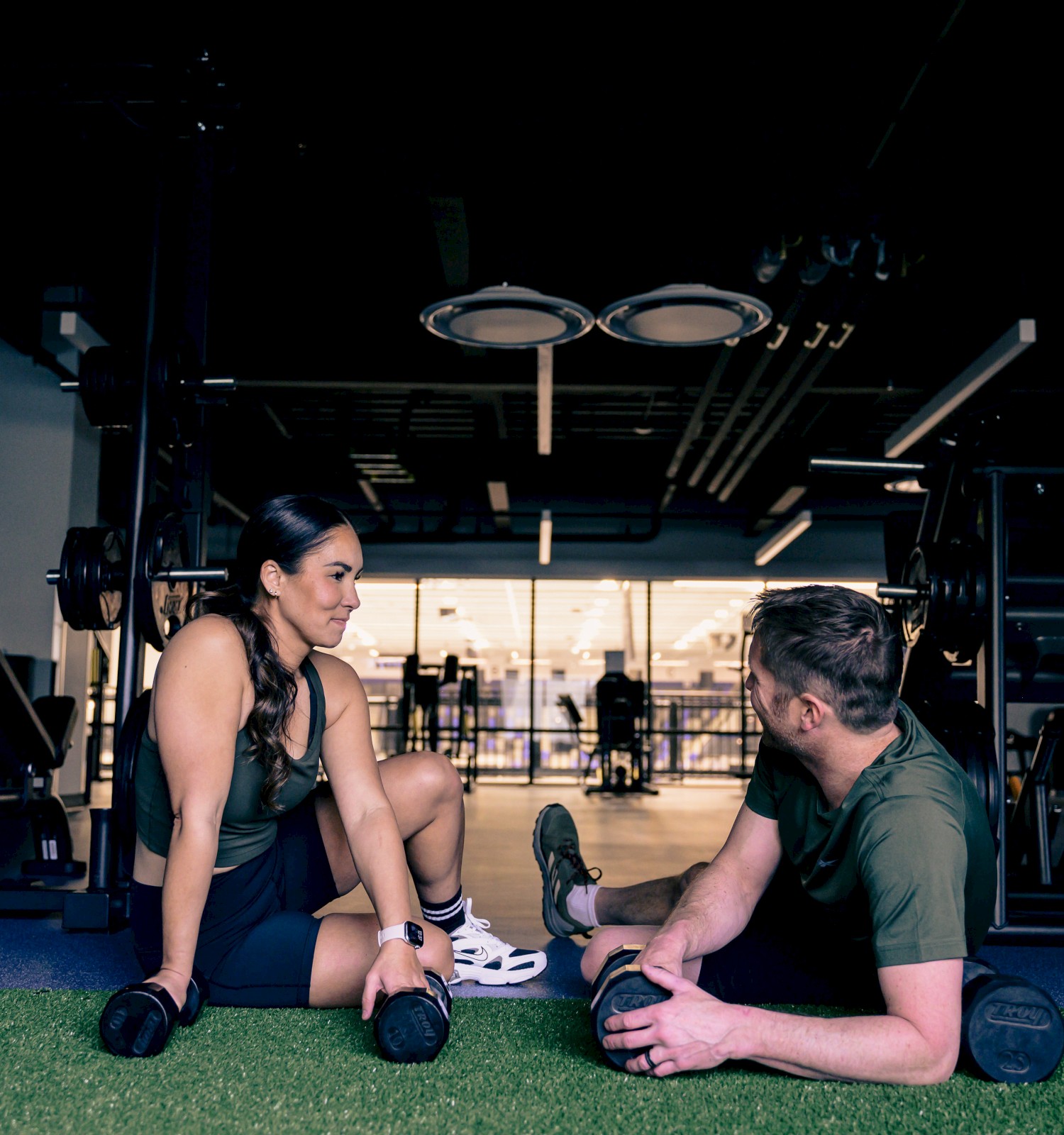 Two people are having a workout break on the gym floor, with weights and fitness equipment around them.