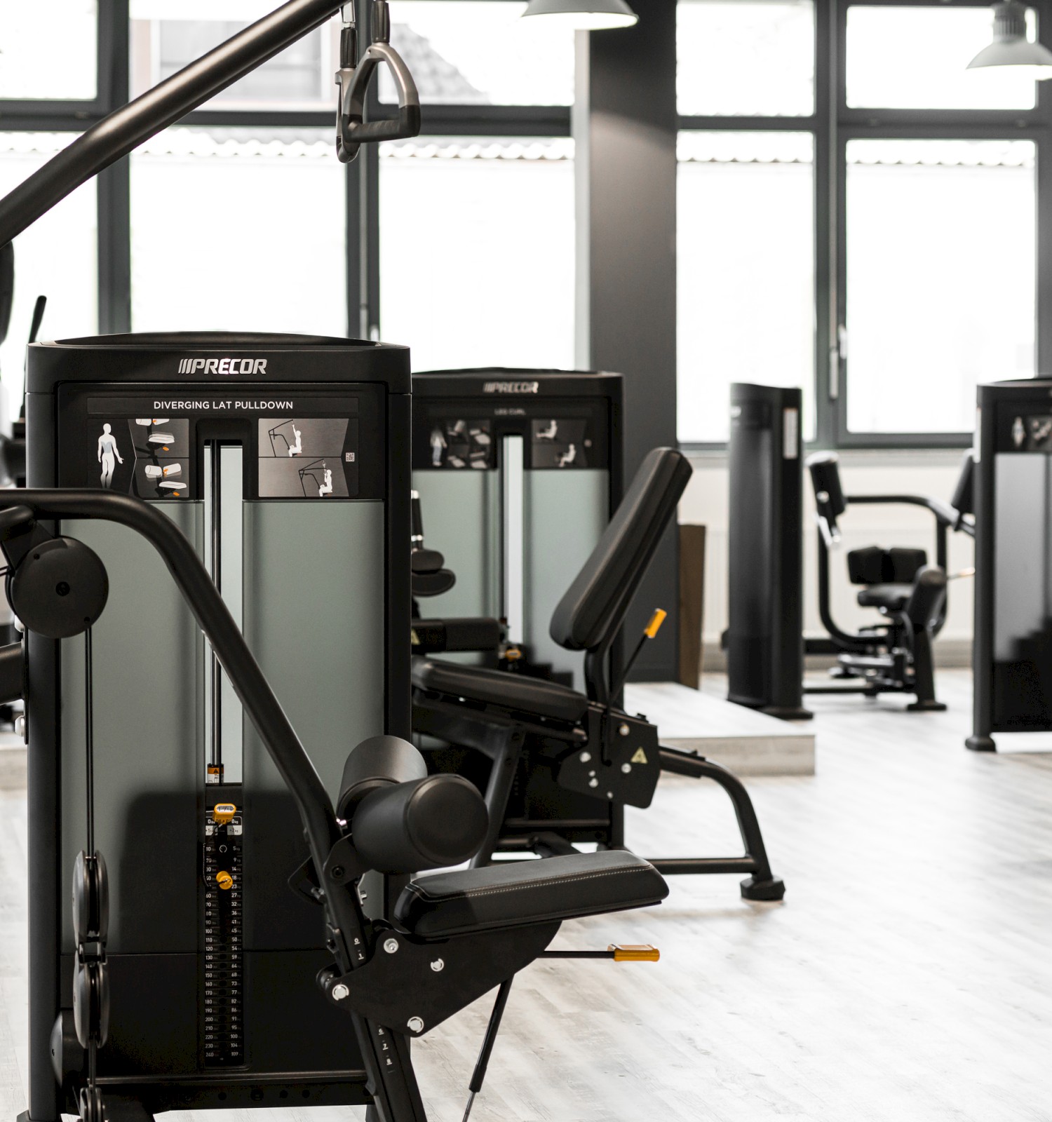 The image shows a modern gym interior with various exercise machines, including strength-training equipment, set against large windows.