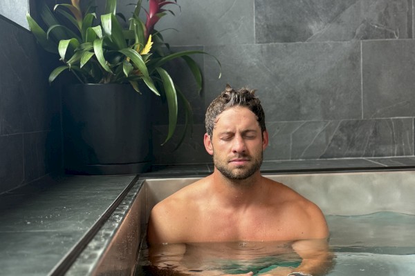 A person is relaxing in a modern indoor bathtub with plants nearby, surrounded by grey tiled walls.