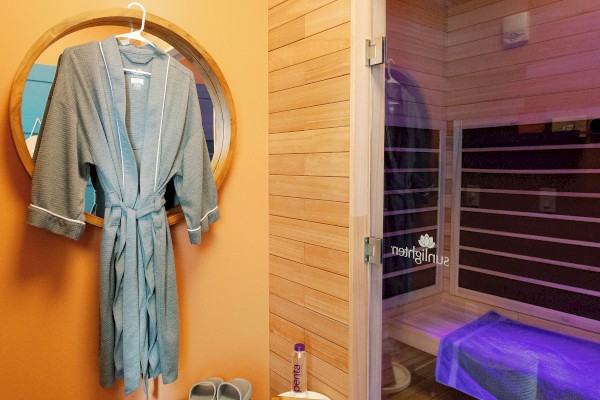 The image shows a spa room with a robe, slippers, bench, and an infrared sauna featuring purple lighting behind a glass door.