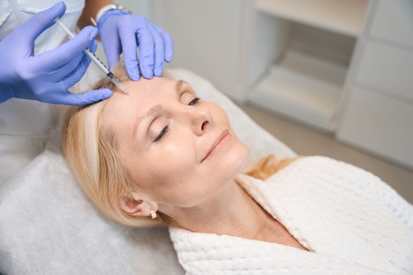 A person receiving a cosmetic injection on their forehead by a gloved professional, lying relaxed in a clinical setting.