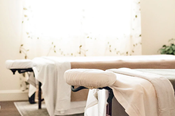 A serene room with massage tables, white linens, and soft sunlight through a window, creating a relaxing atmosphere.