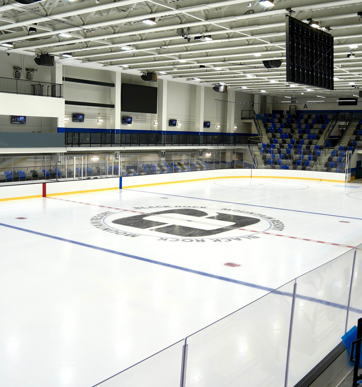 An empty ice hockey rink with seating, glass boards, and a central logo on the ice, under bright arena lights.