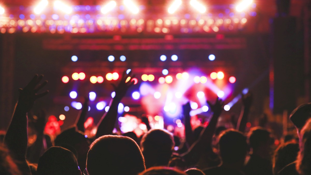 A crowd is enjoying a concert with colorful stage lights and people raising their hands in the air.
