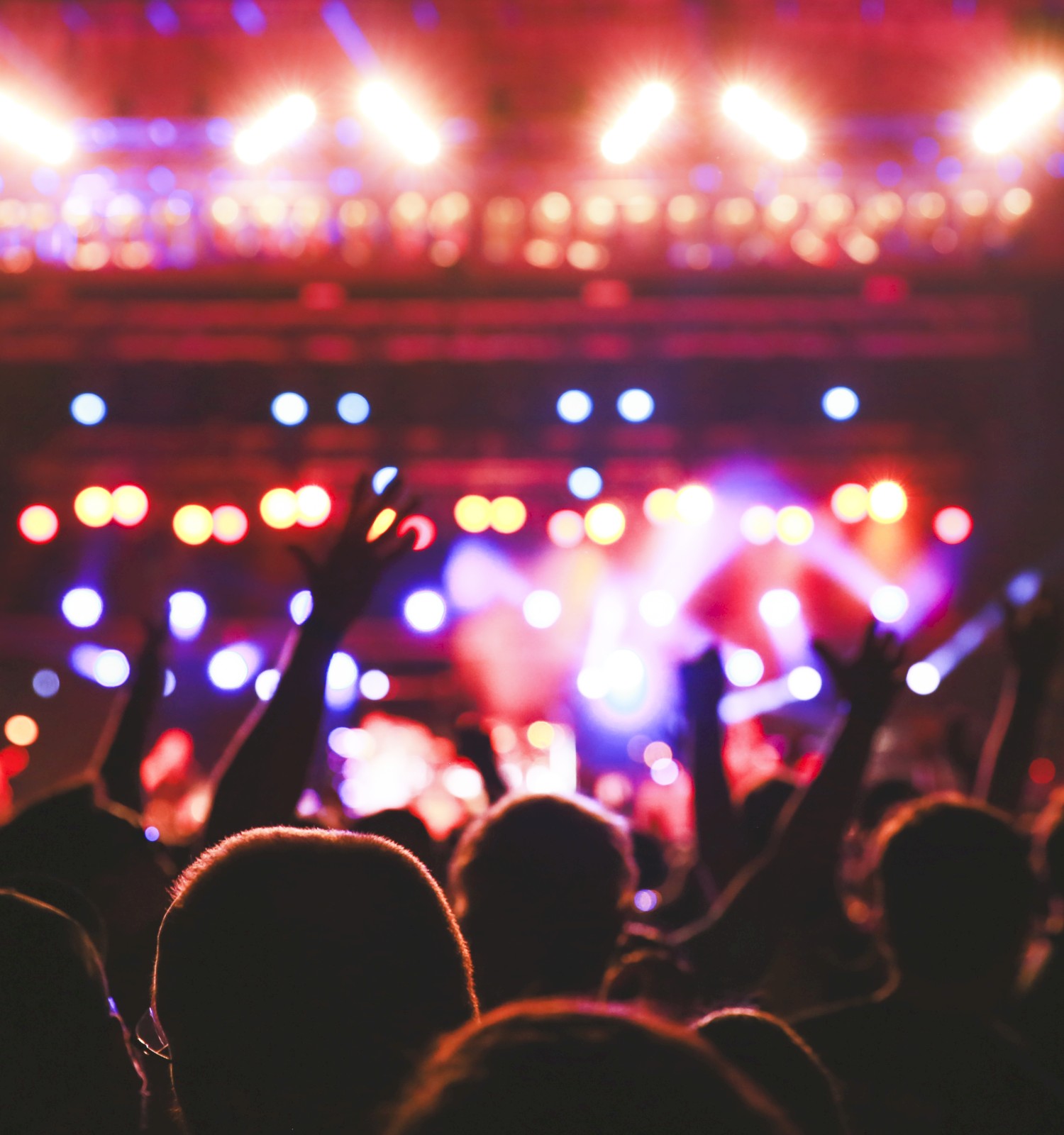 A crowd is enjoying a concert with colorful stage lights and people raising their hands in the air.