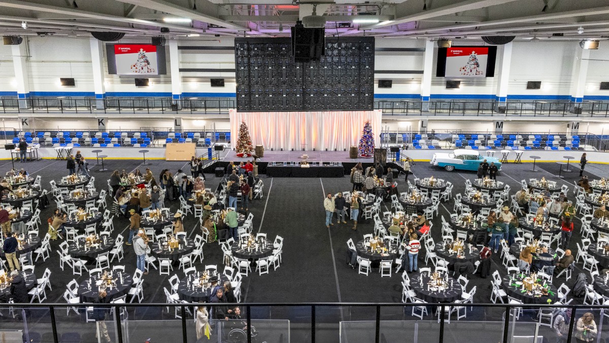A large indoor event hall with many round tables and chairs arranged for a banquet, a stage with curtains, and people mingling in groups.