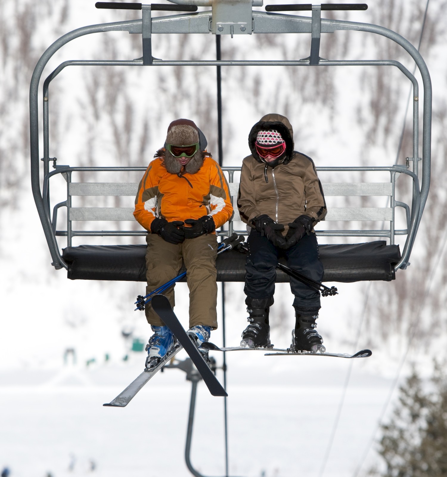 Two people on a ski lift, dressed in winter gear, with snowy trees and a mountain background.