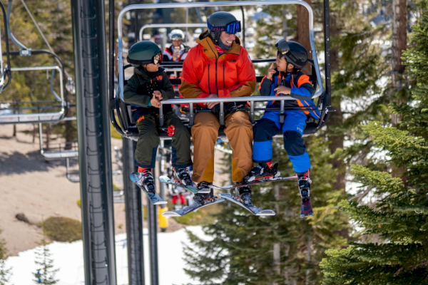 Three people in ski gear sitting on a ski lift, surrounded by snow and trees, heading up the mountain.
